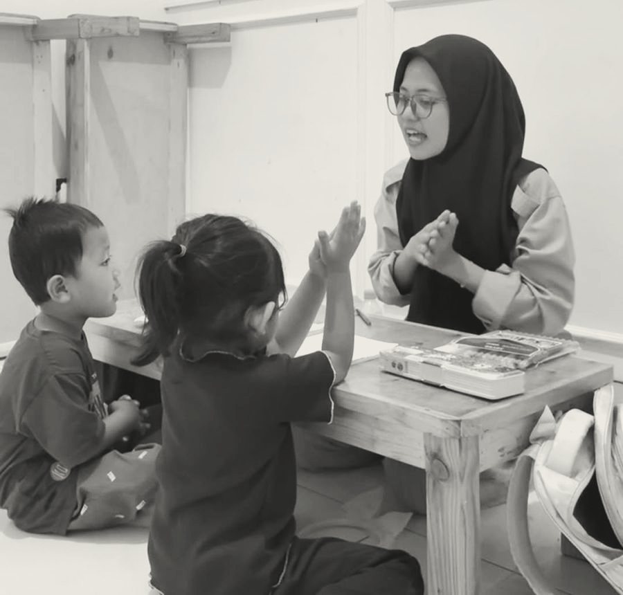 Seorang guru Lilago berkerudung bertepuk tangan sambil bernyanyi bersama dua anak kecil di meja belajar rendah.