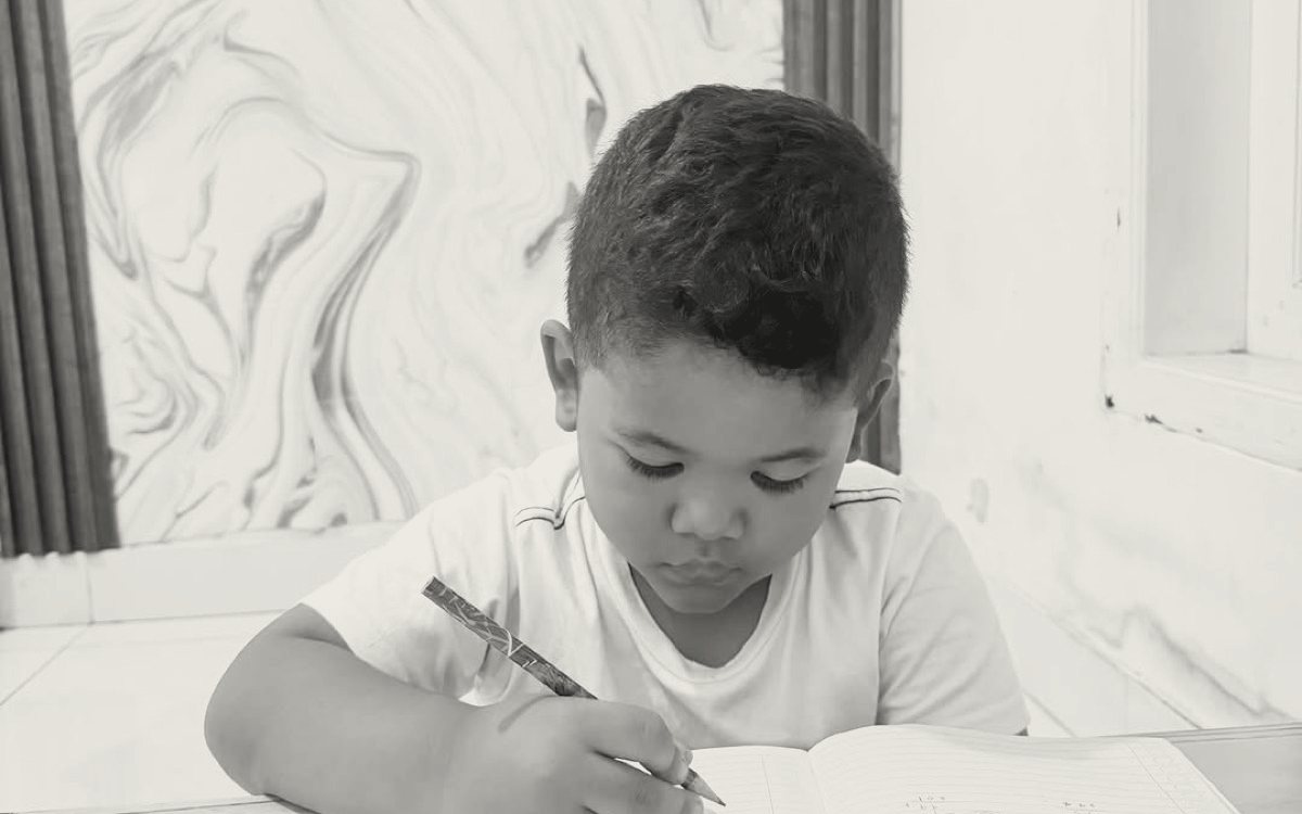 Anak laki-laki berkaos putih mengerjakan soal penjumlahan di buku tulis bergaris dengan pensil merah, di meja kayu ruang belajar Lilago.