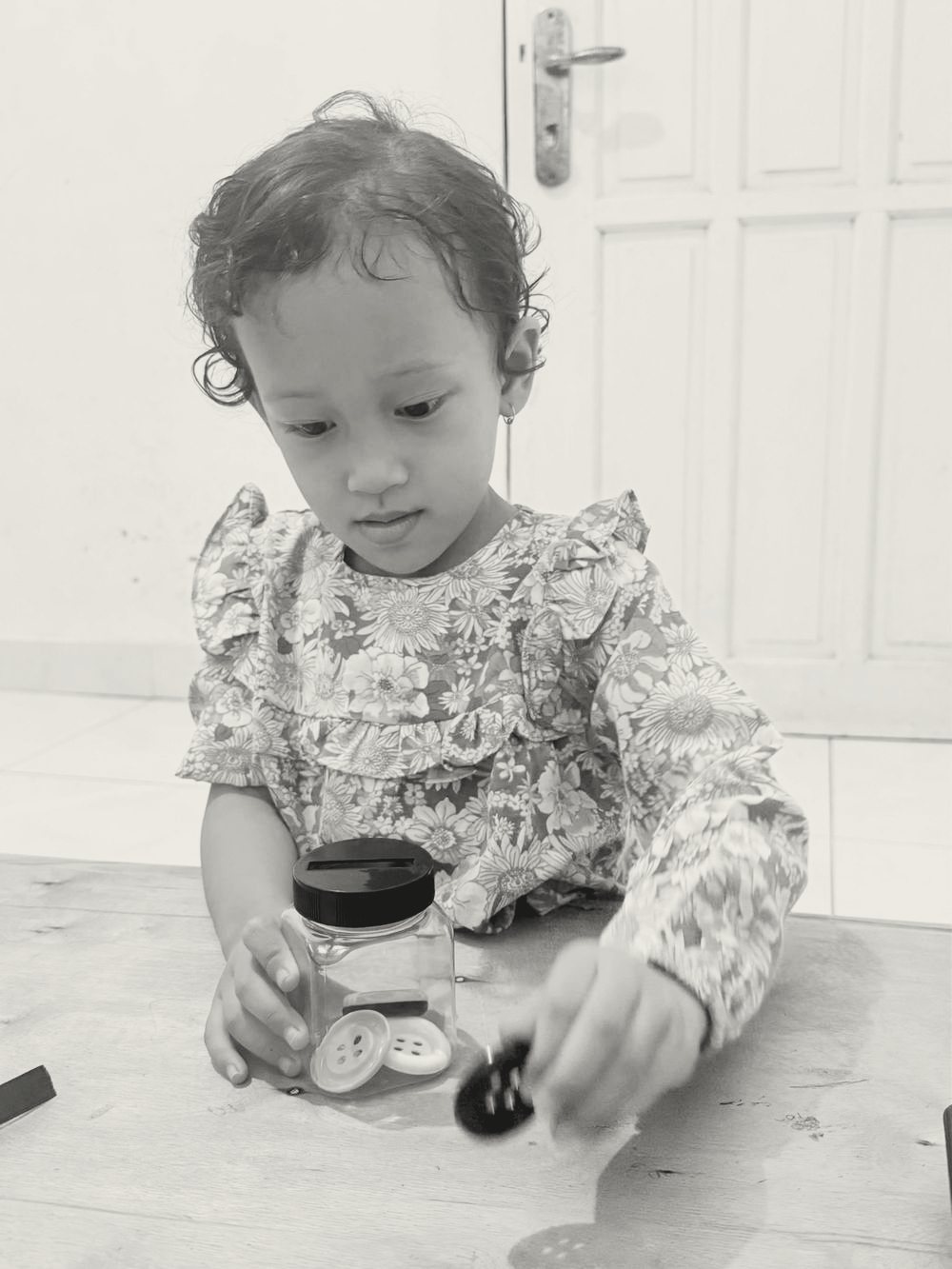 Anak perempuan yang sama memasukkan kancing baju satu per satu ke dalam toples kecil.