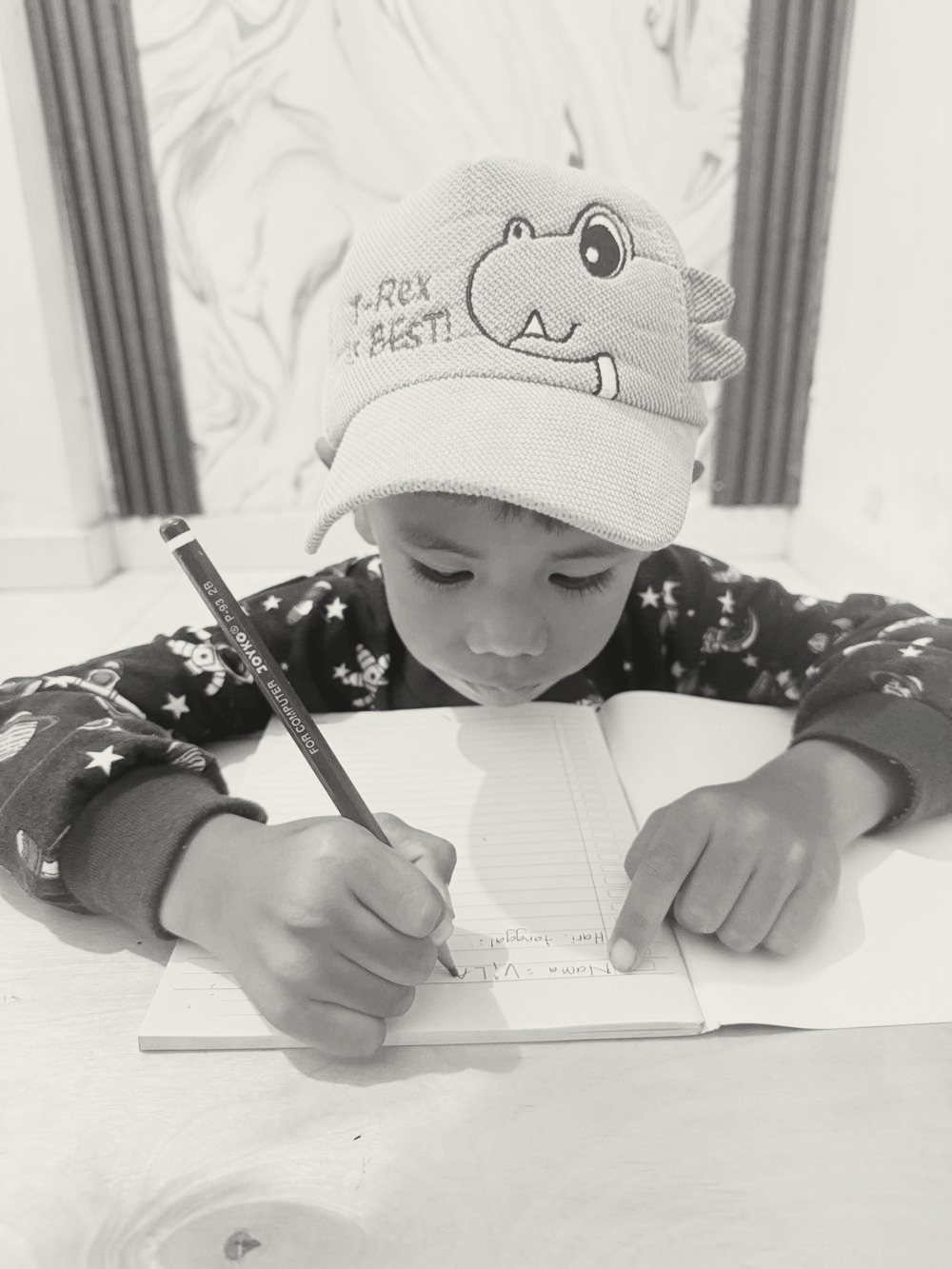 Anak laki-laki bertopi dinosaurus menulis namanya di buku tulis sambil menunjuk baris dengan jarinya.