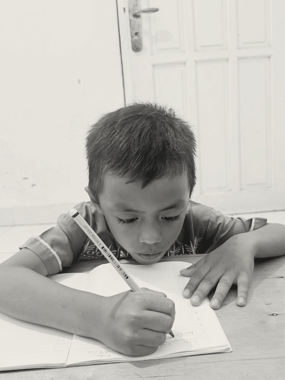 Anak laki-laki berbaju batik cokelat menulis dengan pensil kuning, kepala hampir menempel ke buku.