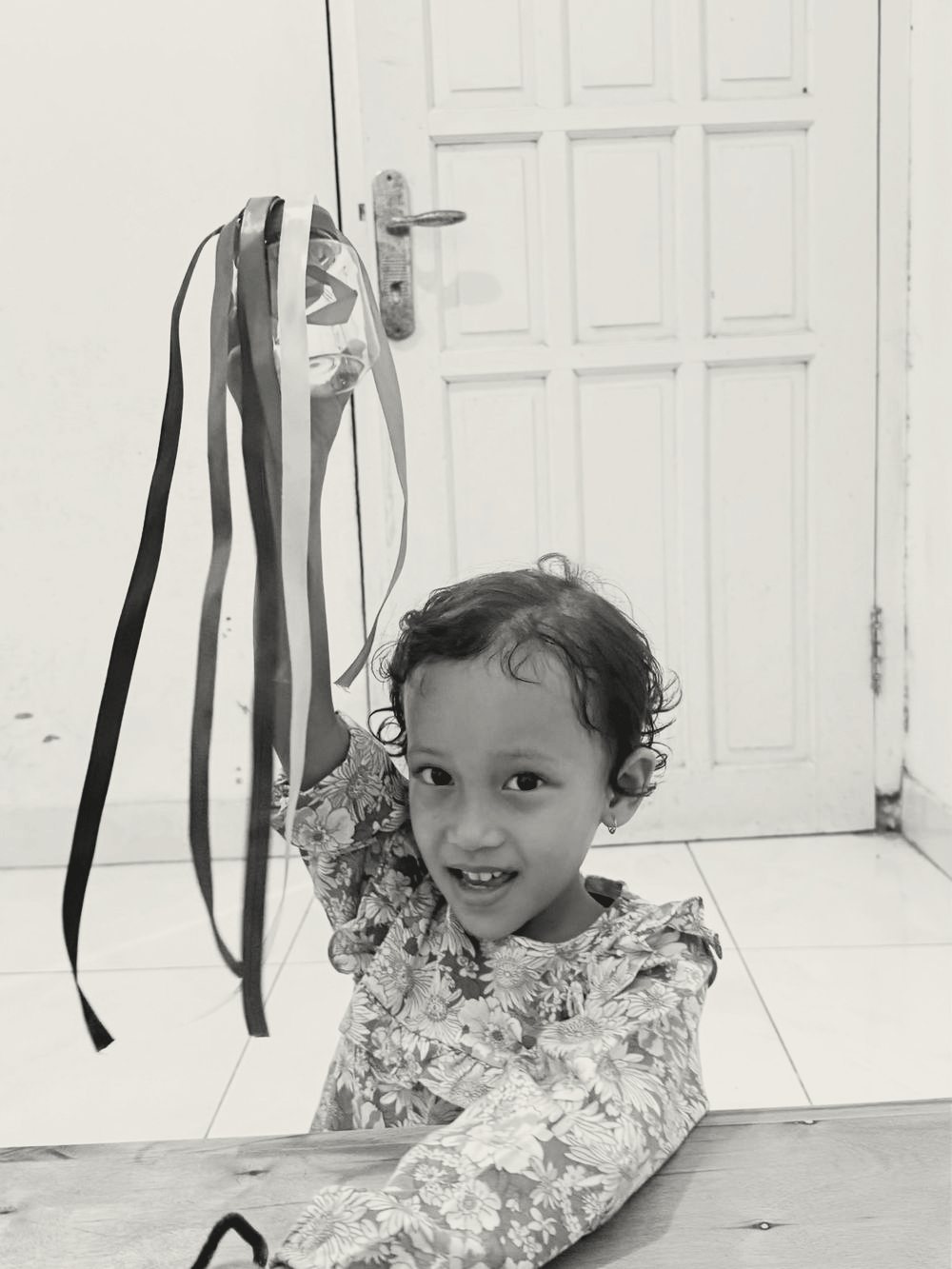 Anak perempuan kecil berbaju motif bunga tersenyum sambil mengangkat botol berpita warna-warni.