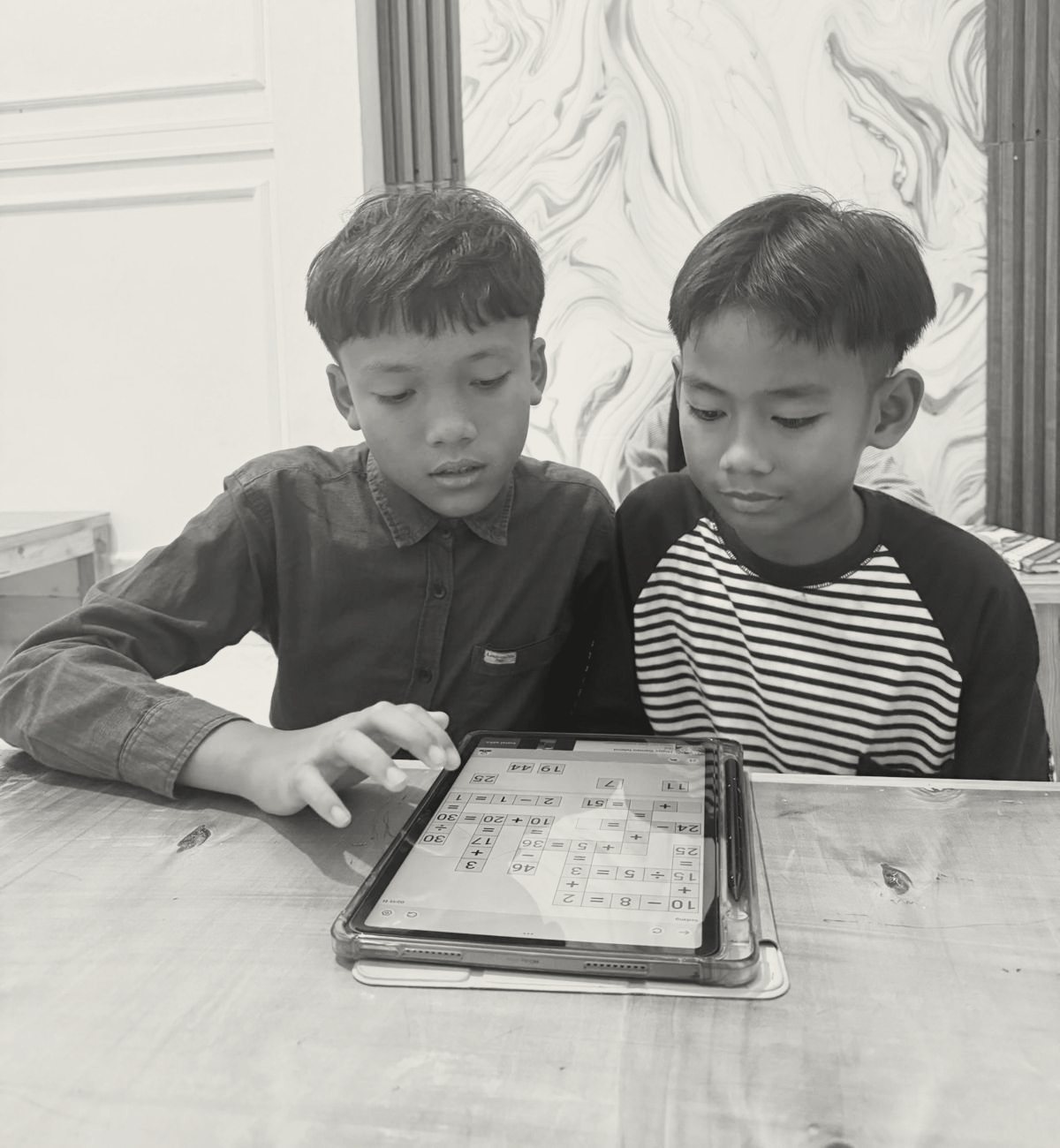 Dua anak laki-laki duduk berdampingan mengerjakan teka-teki angka di tablet.